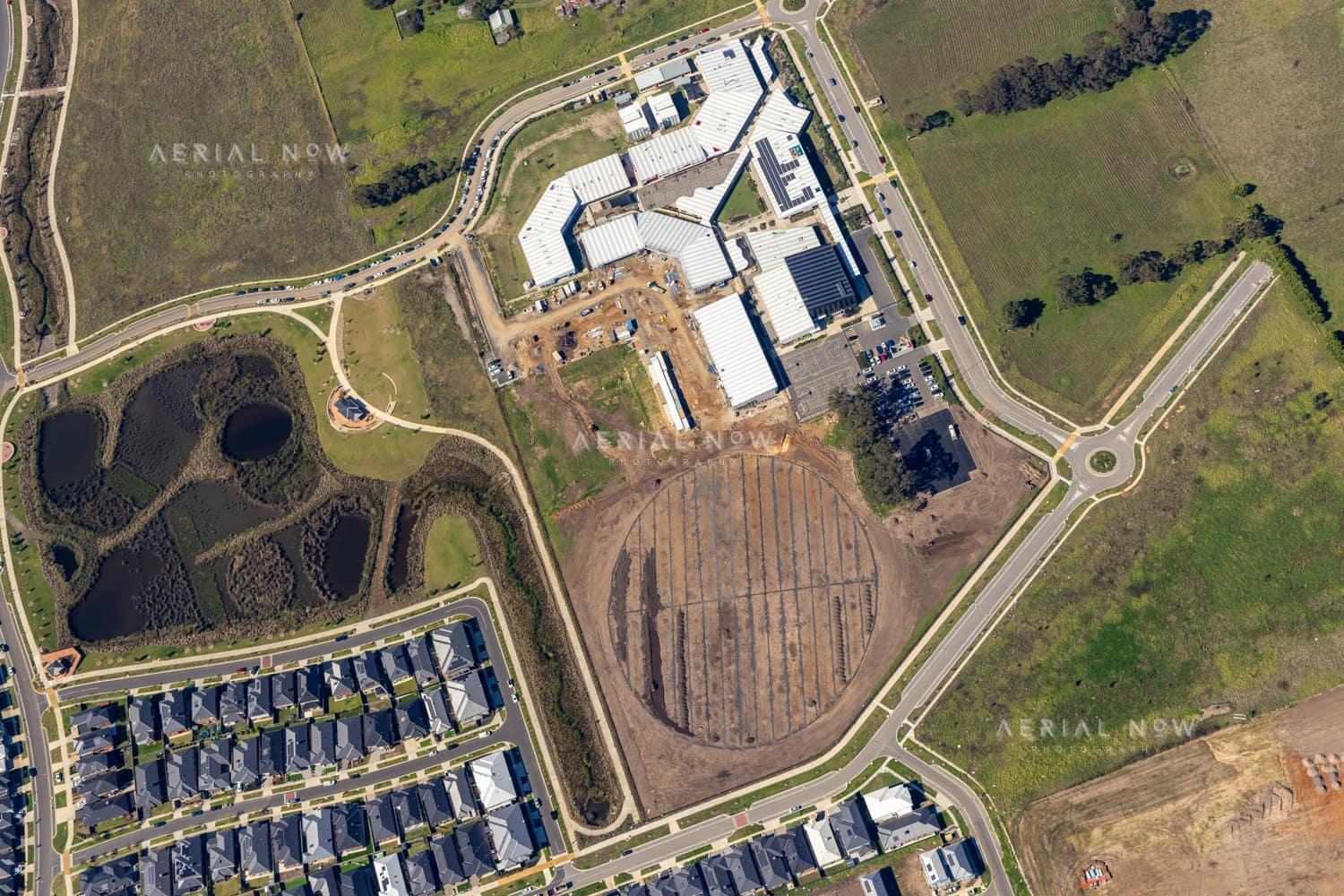 Aerial view of Clyde Secondary College on Woodfield Avenue in Clyde, showing campus buildings, hardcourts, car parking, earthworks and a prepared sports oval area beside nearby residential streets.