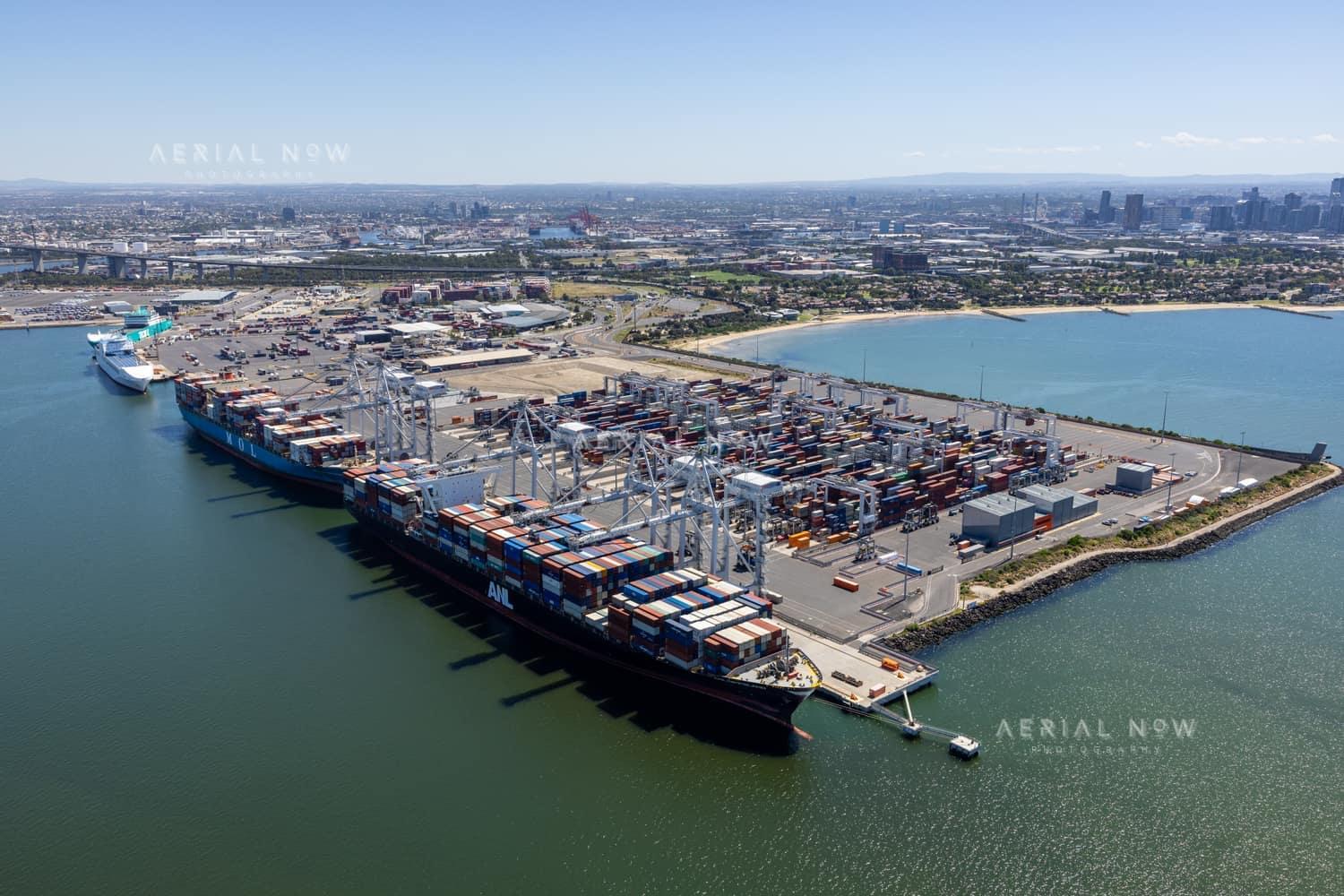 Aerial view of container ships berthed at Webb Dock East in Port Melbourne, with ship-to-shore cranes, container stacks, Port Phillip Bay and the Melbourne CBD skyline in the background.
