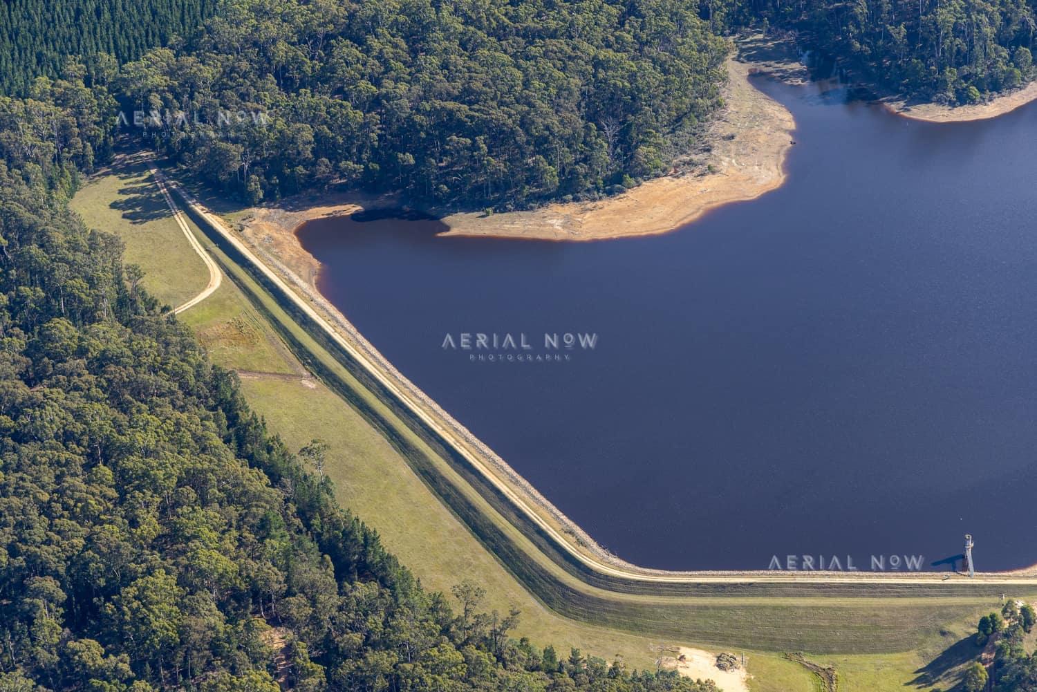 Aerial view of Bostock Reservoir at Spargo Creek, showing the long grassed dam crest, dark reservoir water, exposed shoreline and surrounding forest.