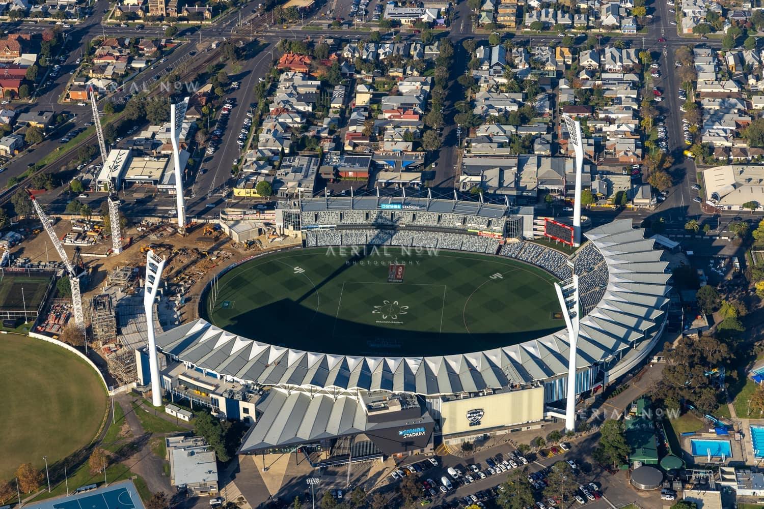 Aerial view of GMHBA Stadium at Kardinia Park in South Geelong, showing the oval, seating bowl, light towers and a construction zone with cranes beside one stand.