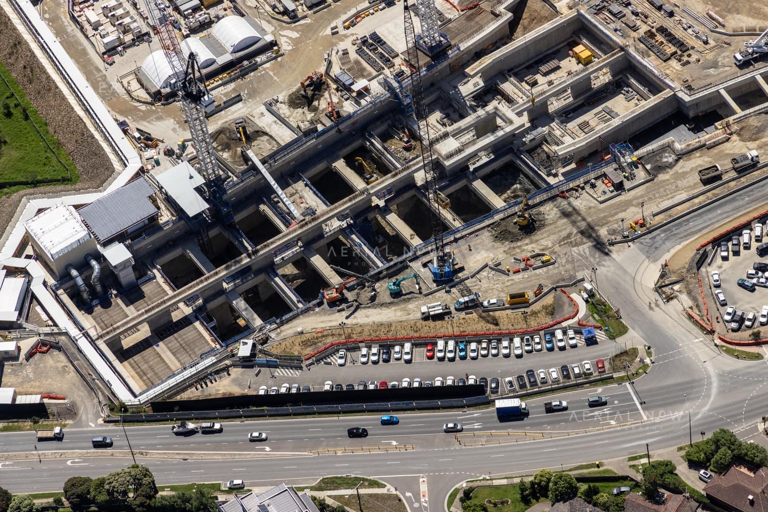 Aerial view of North East Link construction works in Bulleen, showing concrete tunnel and road structures, cranes, work zones, parked vehicles and traffic on the adjoining arterial road.