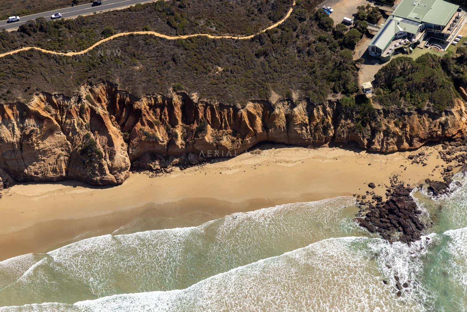 Aerial view of Demons Bluff Beach at Anglesea, showing sandstone cliffs, a narrow sandy beach, rocky shoreline, surf and clifftop houses beside a coastal road.