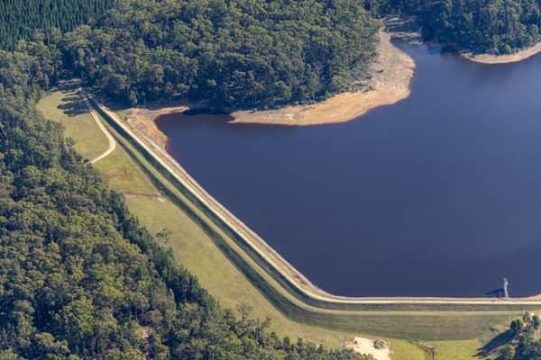 Aerial view of Bostock Reservoir at Spargo Creek, showing the long grassed dam crest, dark reservoir water, exposed shoreline and surrounding forest.