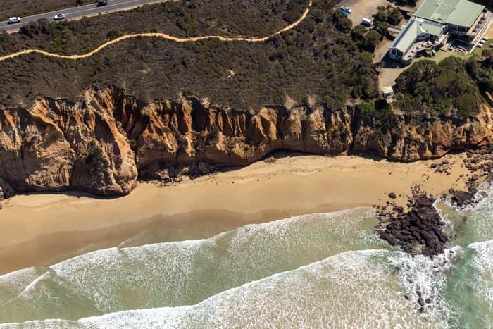 Aerial view of Demons Bluff Beach at Anglesea, showing sandstone cliffs, a narrow sandy beach, rocky shoreline, surf and clifftop houses beside a coastal road.