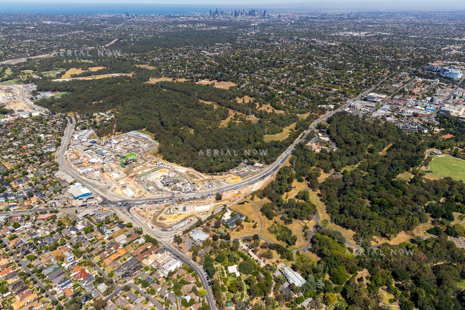 Aerial view of North East Link construction works in Bulleen, with roadworks, surrounding suburban streets, parkland and the Melbourne skyline in the distance.
