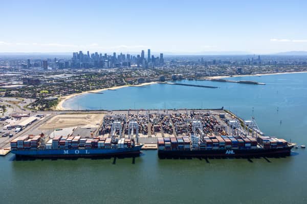 Aerial view of Webb Dock East at the Port of Melbourne, showing container ships, ship-to-shore cranes, container stacks, Port Phillip Bay and the Melbourne CBD skyline in the background.