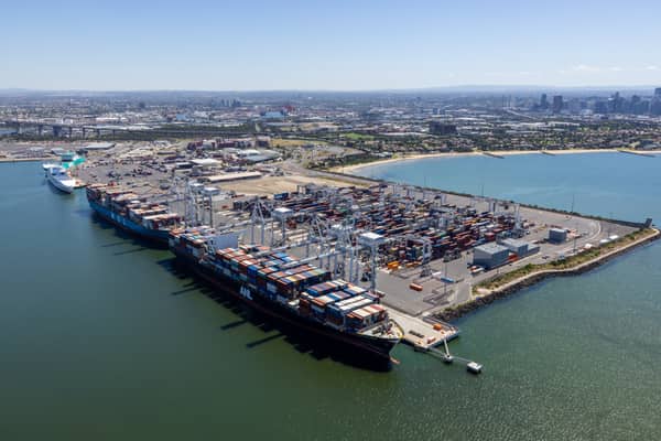 Aerial view of container ships berthed at Webb Dock East in Port Melbourne, with ship-to-shore cranes, container stacks, Port Phillip Bay and the Melbourne CBD skyline in the background.