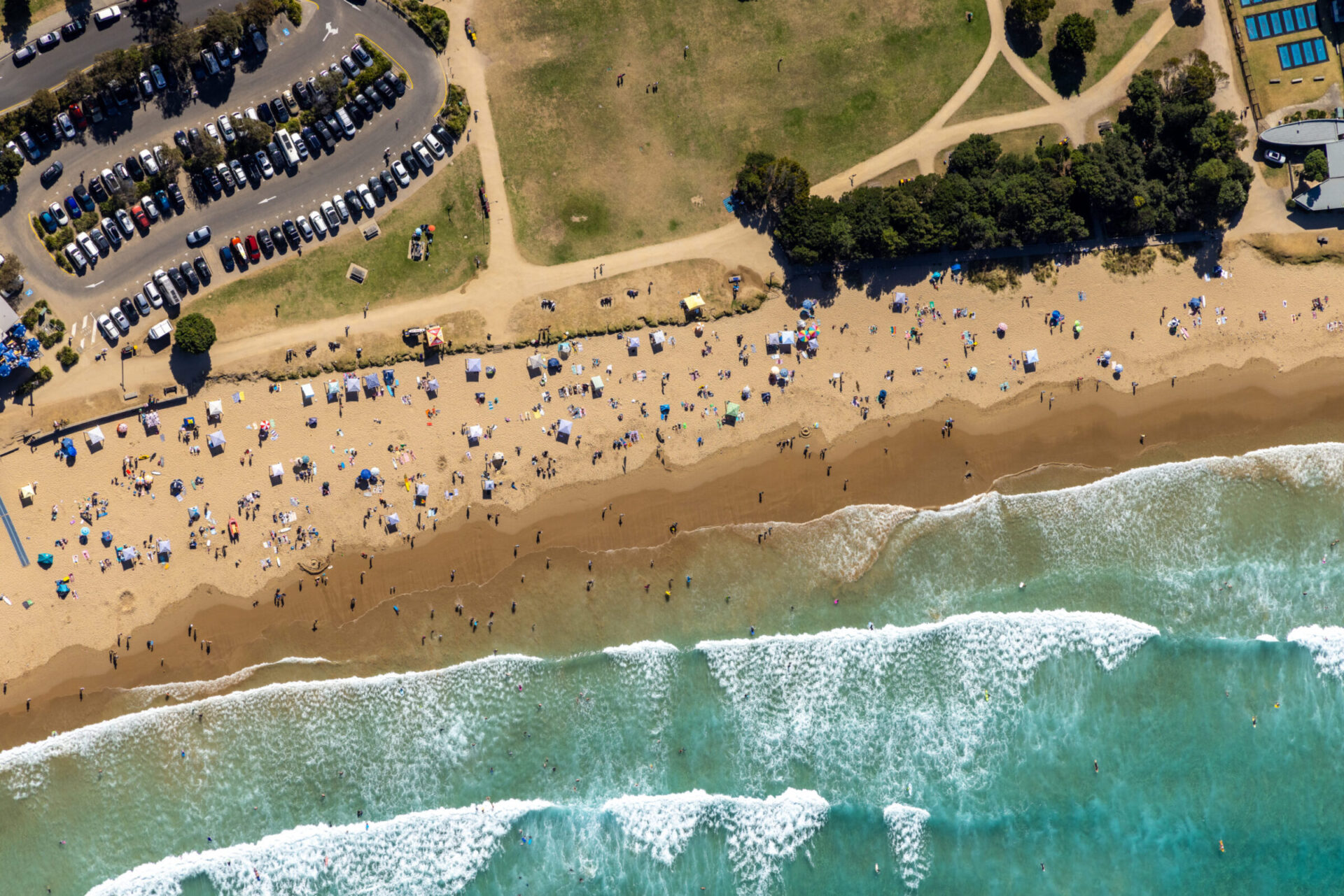 Aerial drone photograph of Lorne Beach on a busy summer day, showing beachgoers, market stalls, surf conditions, foreshore reserve, car park capacity and surrounding parkland within Surf Coast Shire Council's jurisdiction.